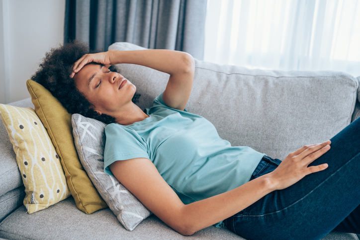 Photo shows woman on couch with hand resting on head and eyes closed